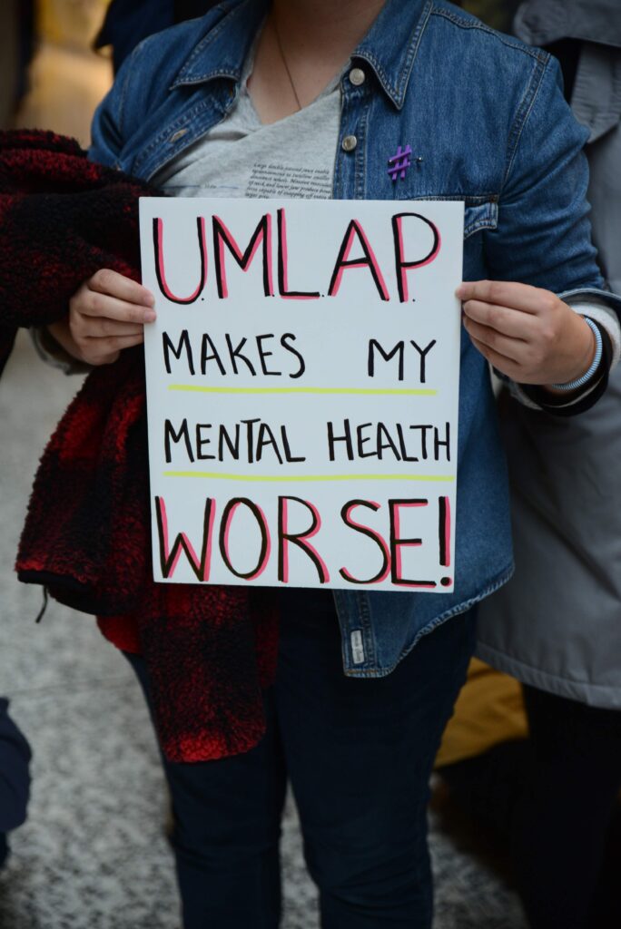 A student holds a sign protesting against the University-Mandated Leave of Absence Policy (UMLAP).